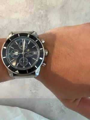 Breitling Superocean Heritage klocka - Snygg Breitling Superocean Heritage kronograf med svart urtavla och datumvisning. Klockan har ett robust mesh-armband i rostfritt stål och tydliga vita markeringar. Klassisk rund form och sportig känsla, perfekt för dig som gillar stilrena accessoarer.