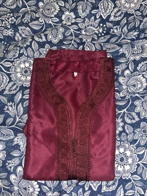 Vinröd kaftan med brodyr - Säljer en vinröd kaftan med dekorativ brodyr längs halsringningen och framtill. Plagget har en klassisk rak form och är tillverkat i ett glansigt tyg som ger en elegant känsla. Perfekt för dig som vill ha något unikt och stilrent.