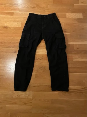 Svarta cargobyxor med fickor - Säljer ett par svarta cargobyxor med raka ben och stora sidofickor på båda benen. Byxorna har klassisk cargomodell med fickor bak och fram, samt bälteshällor i midjan. Perfekta för en avslappnad och trendig streetwear-look.