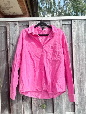 Barbie pink oversized manchesterskjorta i bomull - Supersnygg oversized skjorta i  hot pink. Skjortan har klassisk krage, bröstficka och knappar framtill. Tillverkad i mjuk bomull och har långa ärmar. Perfekt för en avslappnad och trendig look. Enkel att styla! 