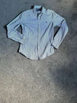 Randig skjorta från Polo Ralph Lauren - Klassisk skjorta från Polo Ralph Lauren i vitt och blått med tunna vertikala ränder. Skjortan har lång ärm, knappar framtill och den ikoniska broderade loggan på bröstet. Tillverkad i mjuk bomull för en bekväm känsla.