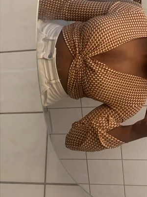 Brunvitrutig crop top med cut-out - Trendig brun och vit crop top med rutigt mönster och cut-out detalj framtill. Toppen har långa ärmar och en åtsittande passform som framhäver midjan. Perfekt för dig som vill sticka ut med en cool och modern stil.