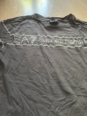 Svart EA7 Emporio Armani t-shirt - Snygg svart t-shirt från EA7 Emporio Armani med vit logga och text över bröstet. Klassisk passform och rund halsringning. Perfekt till jeans eller shorts för en clean och stilren look.