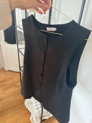 Svart väst med knappar från Stockh LM - Stilren svart väst från Stockh LM med rund halsringning och klädda knappar framtill. Västen har en rak passform och är tillverkad i ett slätt material som ger en clean look. Perfekt att styla över en skjorta eller t-shirt.🖤