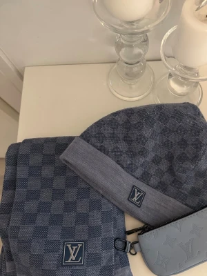 Is blå LVuitton set - Snygg blå halsduk från Louis Vuitton med klassiskt rutmönster och diskret LV-logga. Halsduken är stickad i mjukt material och har en modern, stilren look som passar perfekt till höst och vinter. Matcha med mössa för en komplett outfit. Vinter säsongen är här snart. Matchar med allt.