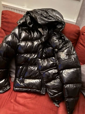 Moncler Maya - Säljer en svart, glansig dunjacka från Moncler med stor huva och klassisk Moncler-logga på ärmen. Jackan har quiltad design, dragkedja framtill och praktiska fickor. Perfekt för kalla vinterdagar och riktigt snygg streetstil.