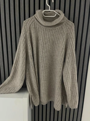 Beige polotröja från 157 - Mysig beige stickad polotröja från 157 med ribbad struktur. Tröjan har en hög polokrage, långa ärmar och en avslappnad passform. Perfekt för kalla dagar och enkel att styla med jeans eller kjol.