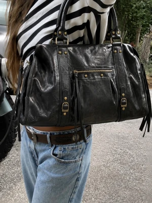 Svart handväska med nitar och dragkedja - Äkta läder ”made in Italy” Snygg svart handväska i skinnimitation med detaljer i guld, dragkedja framtill och dubbla handtag. Väskan har dekorativa nitar och spännen samt fransdetaljer på sidorna. Perfekt för dig som gillar en edgy och trendig look.