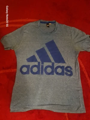Blå Adidas t-shirt med stor logga - Säljer en blåmelerad t-shirt från Adidas med stor blå logga på bröstet. Klassisk rund hals och korta ärmar. Perfekt för dig som gillar sportig och avslappnad stil. Tillverkad i mjuk bomull för skön känsla hela dagen.