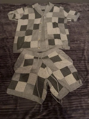 Grått stickat set med rutmönster - Säljer ett snyggt tvådelat set med kortärmad skjorta och shorts i stickat material. Setet har ett rutigt mönster i olika nyanser av grått och vitt, med knappar framtill på skjortan och dragsko i midjan på shortsen. Perfekt för dig som gillar en avslappnad men trendig stil.