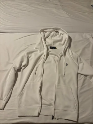 Vit hoodie från Polo Ralph Lauren - Snygg vit hoodie från Polo Ralph Lauren med dragkedja, två fickor framtill och klassisk huva med dragsko. Ikoniska Polo-loggan broderad i svart på bröstet. Tillverkad i mjukt material som känns skönt mot huden. Perfekt för en clean och stilren look.