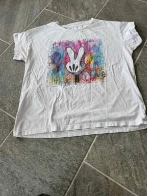 Vit t-shirt med färgglatt tryck - Vit t-shirt med korta ärmar och ett stort färgglatt tryck framtill som föreställer en hand i peace-tecken. Trycket har graffiti-inspirerade färger som rosa, blå, gul och lila. T-shirten har en avslappnad passform och är gjord i mjuk bomull. Ingen storleks lapp finns men skulle uppskatta storleken som en Medium. 