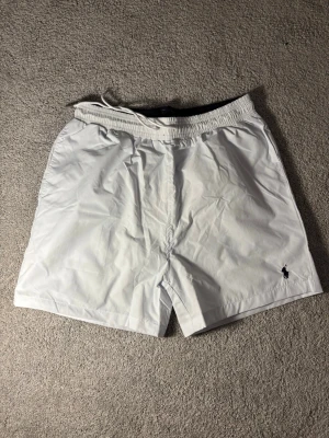 Vita badbyxor från Polo Ralph Lauren - Snygga vita badbyxor från Polo Ralph Lauren med elastisk midja och dragsko. Klassisk passform och diskret logga broderad i svart på vänster ben. Tillverkade i snabbtorkande material, perfekta för stranden eller poolen.