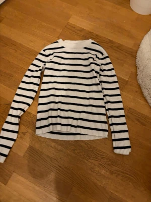 Vit stickad tröja med svarta ränder - Säljer en ribbstickad vit tröja med svarta horisontella ränder. Tröjan har långa ärmar och rund halsringning. Perfekt för dig som gillar klassisk och enkel stil med en twist.