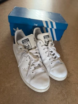 Adidas Stan Smith vita sneakers - Klassiska vita Adidas Stan Smith sneakers med lågt skaft och svarta detaljer på hälen. Skorna har perforerade ränder på sidorna, vit sula och ikonisk Stan Smith-logga på plösen. Tillverkade i läder med textilfoder och snörning framtill.