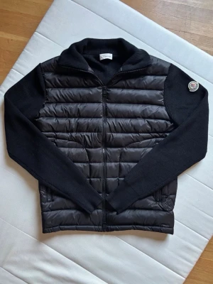 Svart dunväst från Moncler - Snygg svart dunväst från Moncler med stickade ärmar och vadderad front. Västen har dragkedja framtill, två fickor och en klassisk Moncler-logga på ärmen. Perfekt för lager-på-lager och ger en clean look till din outfit.