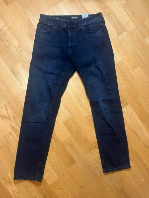 Mörkblå tapered jeans Jack & Jones - Säljer dessa snygga och klassiska mörkblåa jeans från Jack & Jones i storlek w29/L32 med tapered fit och normal midja. Klassisk femficksmodell med knappgylf och smalare ben. Perfekta för dig som gillar en stilren och enkel look. Vid frågor, extra bilder eller liknande är det bara att kontakta mig!