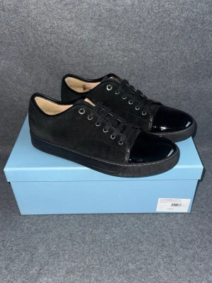 Svarta sneakers från Lanvin - Snygga svarta sneakers i mocka med glansig lackad tå och matchande svarta skosnören från Lanvin. Skorna har en låg profil och svart sula, vilket ger en stilren och modern look. Perfekta för dig som gillar minimalistisk design med en twist.