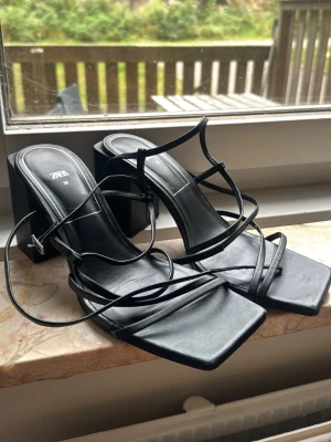 Svarta strappy sandaler från Zara - Snygga svarta sandaler från Zara med tunna remmar och fyrkantig tå. Klackarna är breda och stabila, vilket ger en cool och trendig look. Tillverkade i konstläder med minimalistisk design och justerbara remmar runt ankeln.
