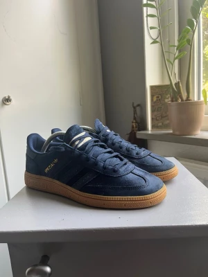 Adidas Spezial blå sneakers 43 1/3 - Säljer ett par Adidas Spezial sneakers i mörkblå mocka med klassiska tre ränder på sidan och gummisula i brunt. Diskret Adidas-logga i guld på sidan och plös. Skorna har en låg profil och tidlös retrostil som passar till det mesta. Lite noppriga skosnören men annars är skorna i bra skick