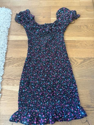 Blommig kortklänning med volang - Supersöt kortklänning med småblommigt mönster i rosa, blått och lila på svart botten. Klänningen har volangdetaljer vid ärmar och nederkant samt en söt knytning framtill. Perfekt för sommarens alla tillfällen!