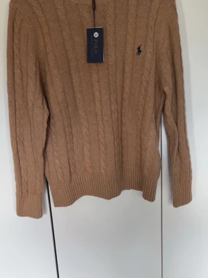 Beige kabelstickad tröja Polo Ralph Lauren - Säljer en beige kabelstickad tröja från Polo Ralph Lauren. Tröjan har rund halsringning, långa ärmar och den klassiska broderade loggan på bröstet. Tillverkad i mjukt material och har ribbade muddar vid ärmslut och nederkant.