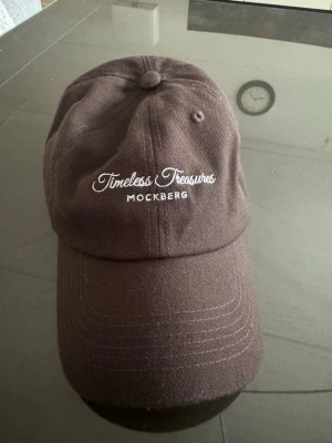 Brun keps från Mockberg - Snygg brun keps från Mockberg med broderad text 'Timeless Treasures' framtill. Kepsen är tillverkad i 80% akryl och 20% ull, vilket ger en mjuk och bekväm känsla. Klassisk böjd skärm och justerbar passform baktill.