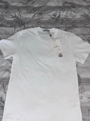 Vit t-shirt från Moncler - Klassisk vit t-shirt från Moncler med rund halsringning och liten Moncler-logga på bröstet. Tillverkad i mjuk bomull och har en clean, stilren look som passar till det mesta. Perfekt basplagg för garderoben.