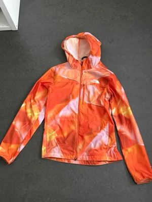 The North Face orange vindjacka  - Orange vindjacka från The North Face med abstrakt mönster i vitt och gult. Jackan har huva, hel dragkedja framtill och två sidofickor. Tillverkad i lätt syntetmaterial och har logga på bröstet och ryggen.