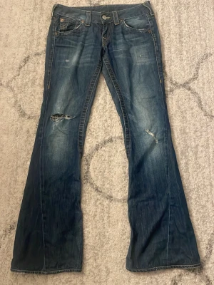 Blå bootcut true religon Jeas - Säljer ett par blå bootcut jeans med slitningar och hål på framsidan. Jeansen har klassiska fem fickor, dekorativa sömmar på bakfickorna och råa kanter vid bensluten. Perfekta för en avslappnad och trendig look.