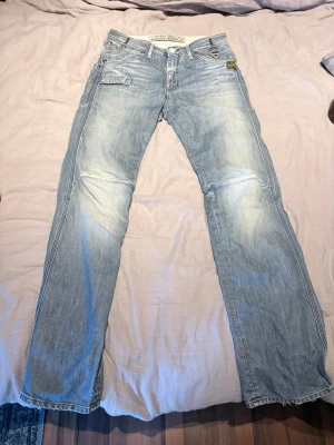 Ljusblå raka jeans med detaljer - Midja 31. Längd 34. Säljer ett par ljusblå G star jeans med raka ben och slitna detaljer. Jeansen har flera fickor, inklusive en extra sidoficka och vita sömmar som sticker ut. Materialet är klassisk denim i bomull och passformen är avslappnad