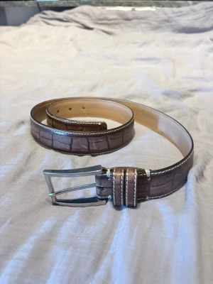 Brunt bälte i skinn med krokopräglat mönster - Snyggt brunt bälte i skinn med krokopräglat mönster och vita kontrastsömmar. Bältet har ett klassiskt silverfärgat spänne och passar perfekt till jeans eller chinos för en stilren look.
