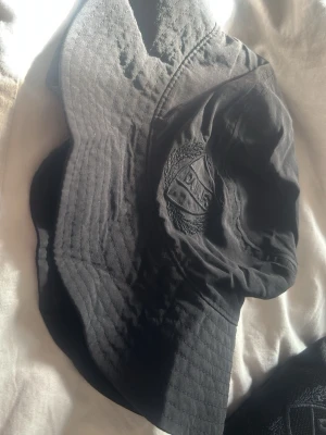 Svart bucket hat med broderad logga - Säljer en svart bucket hat i tyg med broderad logga framtill. Hatten har klassisk form med bred, nedåtlutande brätte och diskret mönster i sömmarna. Perfekt accessoar för en avslappnad streetstil.
