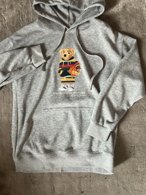 Grå hoodie med Polo Bear-tryck - Säljer en grå hoodie från Ralph Lauren med det ikoniska Polo Bear-trycket på bröstet. Hoodien har huva med dragsko, magficka och ribbade muddar. Perfekt för dig som gillar streetwear och vill ha en chill look.