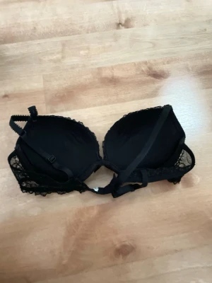 Svart push-up bh med spetsdetaljer - Snygg svart push-up bh med vadderade kupor och fina spetsdetaljer över hela framsidan. Justerbara axelband och hakknäppning bak. Perfekt för dig som vill ha både stöd och en feminin look. Inga etiketter men det är storlek 75A fåen New Yorker. 
