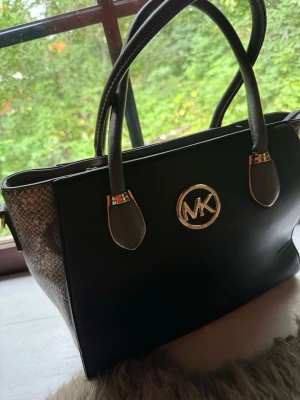 Svart handväska från Michael Kors - Snygg svart handväska från Michael Kors med guldfärgade detaljer och MK-logga framtill. Väskan har två rejäla handtag, ormmönstrade sidor i grått och svart samt dragkedjeficka på baksidan. Perfekt för dig som gillar stilrena accessoarer med en twist.