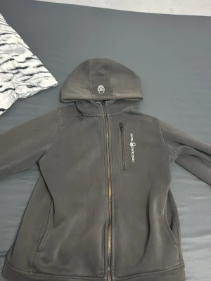 Svart hoodie Sail Racing med dragkedja - Säljer en svart hoodie från Sail Racing med hel dragkedja framtill och en bröstficka med dragkedja. Hoodien har logga på bröstet och på huvan, samt två sidofickor. Tillverkad i mjukt material och har en klassisk passform.