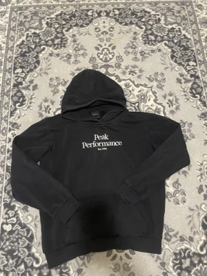 Peak Performance svart tröja  - Snygg svart hoodie från Peak Performance med vit logga och text på bröstet. Klassisk huva och magficka framtill. Tillverkad i mjukt material som känns skönt mot huden. Perfekt för en avslappnad och stilren look.