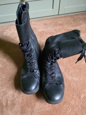 Svarta höga läderboots från Prada - Säljer ett par svarta höga boots från Prada i äkta läder. Skorna har snörning framtill, dragkedja på sidan och en robust gummisula. Klassisk och stilren design med diskret Prada-logga på sidan. Perfekta för dig som vill ha en edgy och exklusiv look. Helt unik modell, köpt på Nitty Gritty. Väldigt mjuka och sköna, tänk på att den går upp en bit på benet och att man behöver en lite vidare byxa för att få i skon. Litet vitt märke på sulan som säkert går att ta bort. Samlarvärde! 