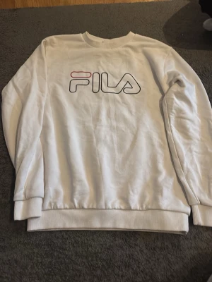Vit sweatshirt från FILA - Säljer en vit sweatshirt från FILA med stor logga framtill i svart och rött. Tröjan har rund halsringning, långa ärmar och ribbade muddar vid ärmslut och nederkant. Perfekt för en avslappnad och sportig stil.