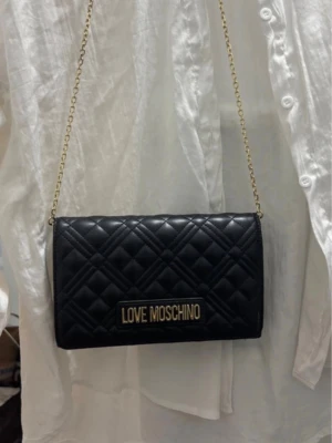 Love Moschino väska  - Jätte fin svart läder väska med guld detalj som kan användas till vardagen eller speciella event.  Jag har knappt använt väskan kanske max 5 gånger dock har jag tappat den och råkar skrapa lädret men det syns knappt. Bandet kan komma av (osäker), och man kan korta bandet för att hänga på axeln. Köptes för ungefär 1300 på zalando. Pris kan diskuteras