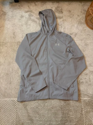 Grå vindjacka från Under Armour - Snygg grå vindjacka från Under Armour med huva och dragkedja framtill. Jackan har långa ärmar, två sidofickor och en stor logga på ryggen. Perfekt för blåsiga dagar och sportiga outfits.