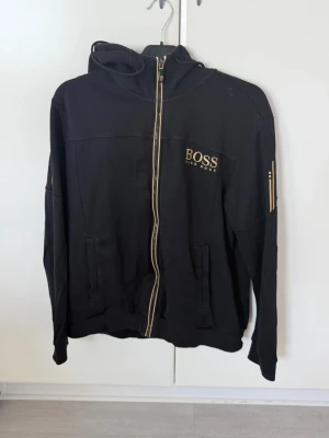 Svart hoodie från BOSS med dragkedja - Snygg svart hoodie från BOSS med guldfärgade detaljer och dragkedja framtill. Hoodien har huva med dragsko, två fickor och BOSS-logga på bröstet. Materialet ser ut att vara mjukt och bekvämt, perfekt för en chill och stilren look.