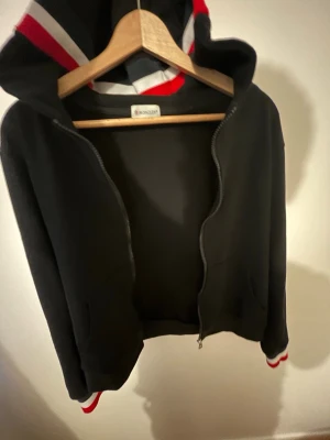Svart hoodie från Moncler med dragkedja - Svart hoodie från Moncler med dragkedja och klassisk Moncler-logga på ärmen. Muddarna har snygga detaljer i rött, vitt och blått. Hoodien har en luva och ribbade muddar vid ärmslut och nederkant. Perfekt för en chill och stilren look.