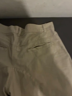 Beige chinos med lejontryck - Snygga beige chinos med raka ben och bälteshällor. Byxorna har två bakfickor och ett coolt vitt lejontryck på ena sidan. Tillverkade i ett mjukt och bekvämt bomullsmaterial som passar perfekt till en avslappnad stil.