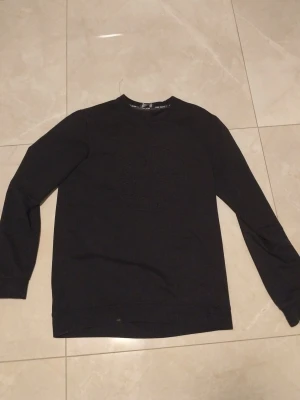 Svart sweatshirt med broderad logga - Snygg svart sweatshirt med rund hals och ribbade muddar. Tröjan har en diskret broderad logga på bröstet och en patch på ena ärmen. Perfekt för en clean och stilren look.