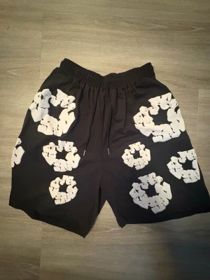 Svarta shorts med vita blommor - Säljer ett par svarta shorts med elastisk midja och snörning. Shortsen har stora vita blommönster över hela plagget och är tillverkade i ett lätt och bekvämt material, perfekt för varma dagar. Passar dig som vill sticka ut med en unik stil.