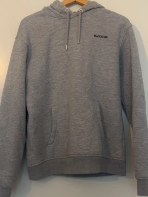 Maggiore hoodie - Säljer en stilren grå hoodie från Maggiore med huva och dragsnören. Hoodien har en klassisk känguruficka framtill och ribbade muddar vid ärmar och nederkant. Perfekt för en chill och avslappnad look. Materialet känns mjukt och bekvämt.