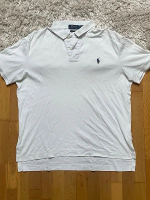 Vit pikétröja från Ralph Lauren - Klassisk vit pikétröja från Ralph Lauren med blå logga broderad på bröstet. Tillverkad i mjuk Pima-bomull och har korta ärmar samt knappar vid halsen. Perfekt för en clean och stilren look. Passar galant som golf tröja. Står M fast passar S