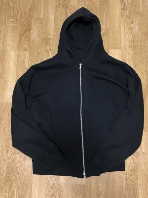 Svart zip hoodie med huva - En klassisk svart hoodie med dragkedja framtill och stor huva. Hoodien har ribbade muddar vid ärmslut och nederkant. Perfekt för en avslappnad och stilren look. Materialet känns mjukt och bekvämt.